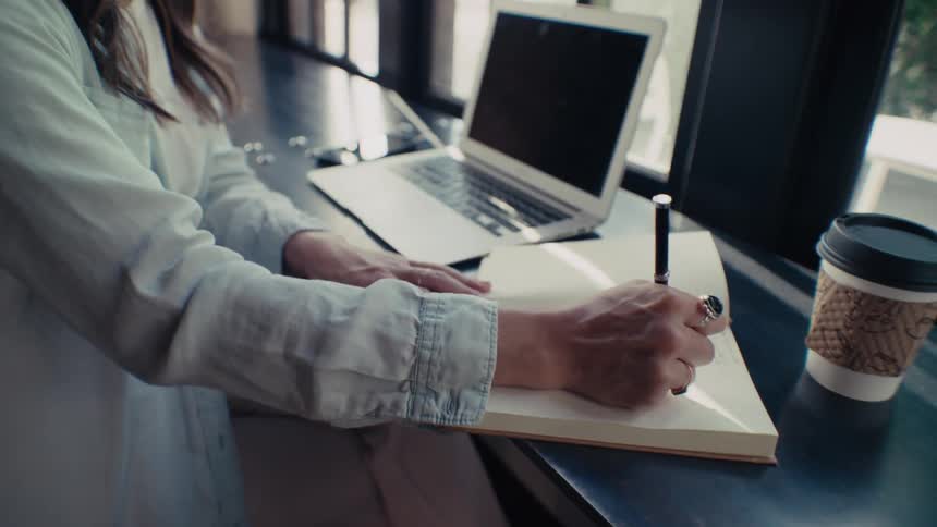 Woman writing in a book while she is sitting at an office desk with an open laptop