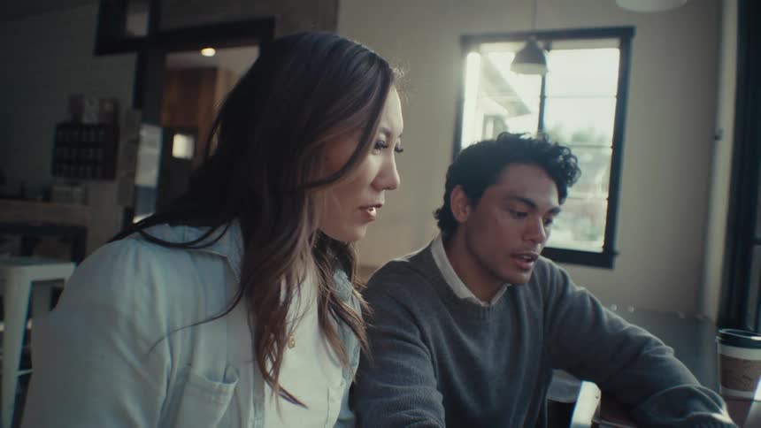 A man and a woman sit side by side at a cafeteria table by a window talking while he types on a laptop