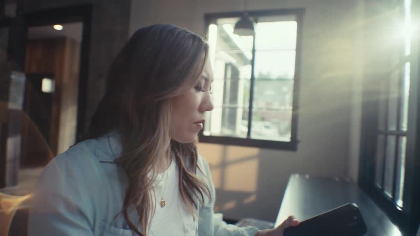 A young woman is holding a book in her hand and looking at a laptop that was just in front of her and then she picks up her cell phone and starts typing on it