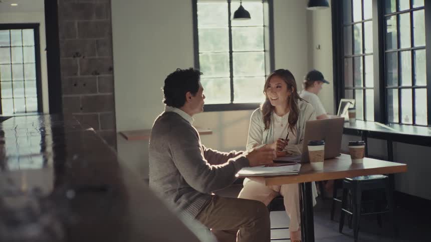 A man and a woman talk and smile at a cafeteria table with a laptop notepads and disposable coffee cups in front of them