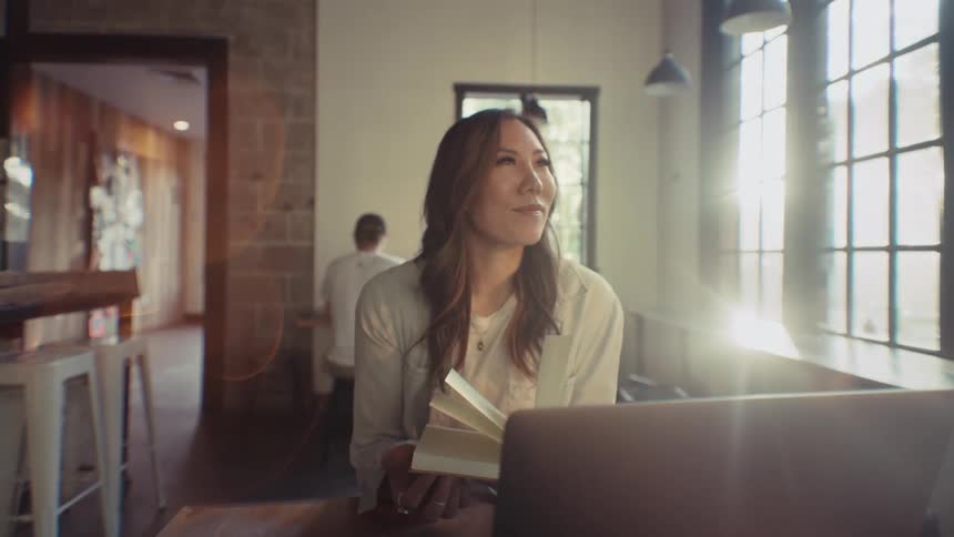 Woman holding a book and being thoughtful with a laptop in front of her