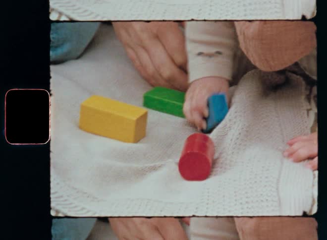 A father or mother tries to help a baby play with colored block toys on a blanket laying outside in the garden or park as the child lets go of the blue wooden building brick before it grabs it again with its tiny and cute hands