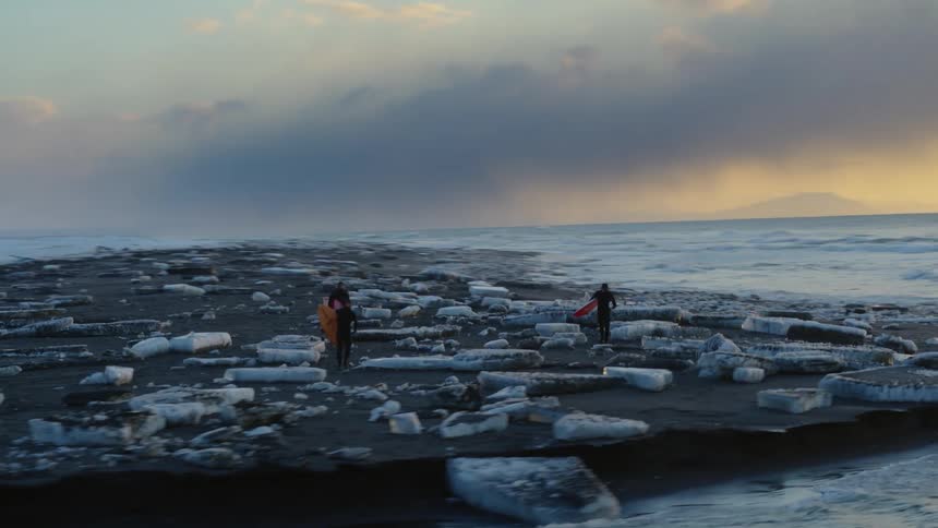 Three people holding surfboards are walking in an area with several pieces of ice on the ground near the sea