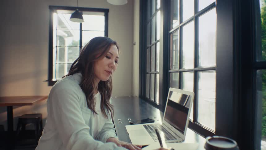 Woman sitting at a table near a large window writing on a notebook near a laptop