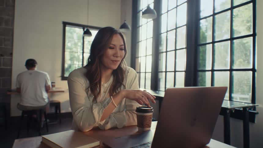 A woman with long hair sitting in a coffee shop reading from a laptop screen