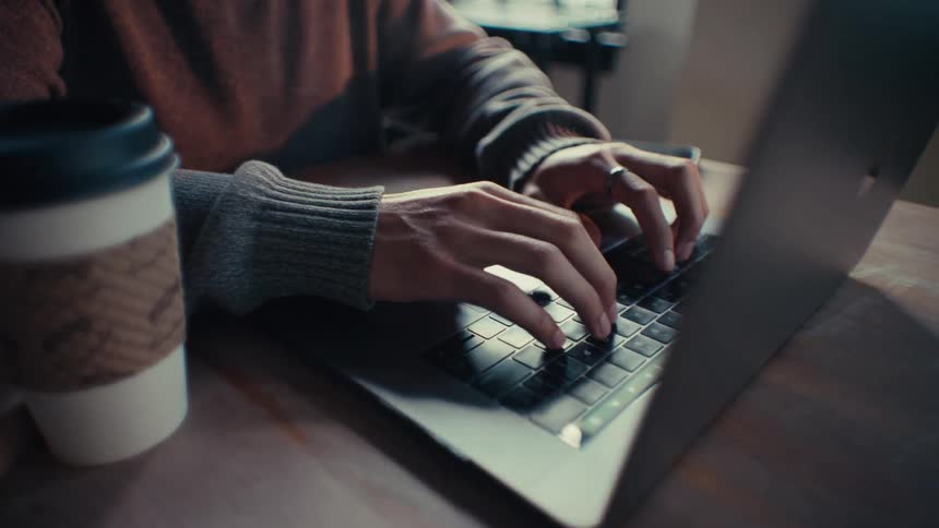 Close up of the hands of a person typing on a laptop keyboard at a wooden table
