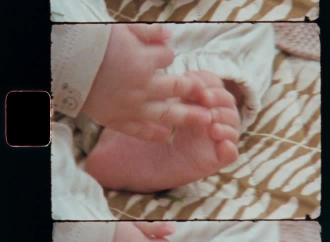 Super 8 mm closeup of a babys feet and hands resting over a blanket near green grass as the small toddler moves around while grabbing his bare feet captured on vintage film with a nostalgic retro vibe