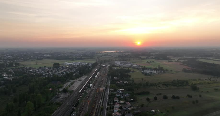 Aerial view of train tracks crossing a pasture field with a city and sunset on the horizon