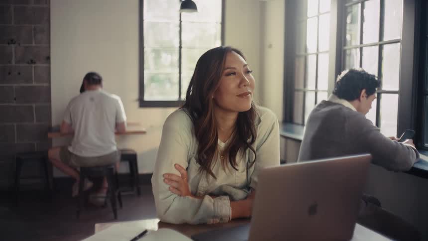 A woman sits in a coffee shop working on a laptop with two other people sitting in the background