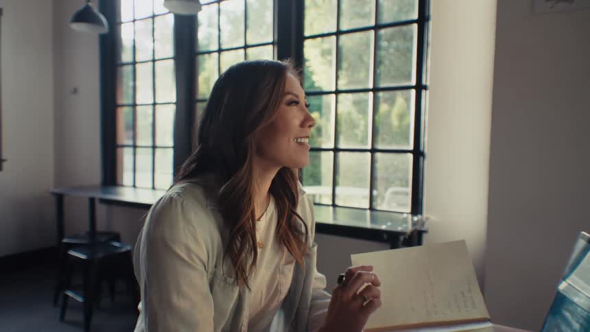 A contemplative woman sitting at a cafeteria table with a laptop in front of her and an open notebook in her hands breaks into a smile