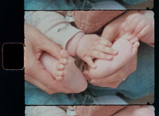 Young caring mother wearing blue jeans sitting outside in the warm sun with baby or toddler on her lap holds the newborn in her hands as the vivid and happy child grabs its tiny feet with curiosity as symbol for parental love and Mother’s Day