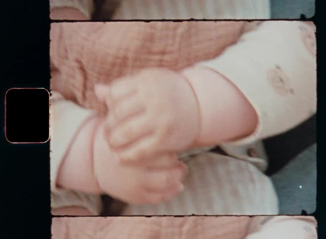 A cute and vivid baby girl or toddler touches or grabs her bare feet with her tiny hands while sitting and playing on her mother’s lap outside during daytime on handheld Super 8 mm close-up symbolizing a happy childhood