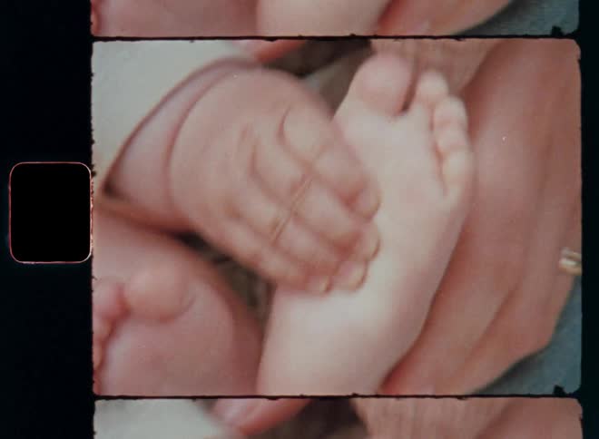 The hands of the adult mother hold the baby’s bare feet as the newborn baby girl sits on her lap touching her soft and pure skin feeling her own body as she is growing up protected by caring and loving parents