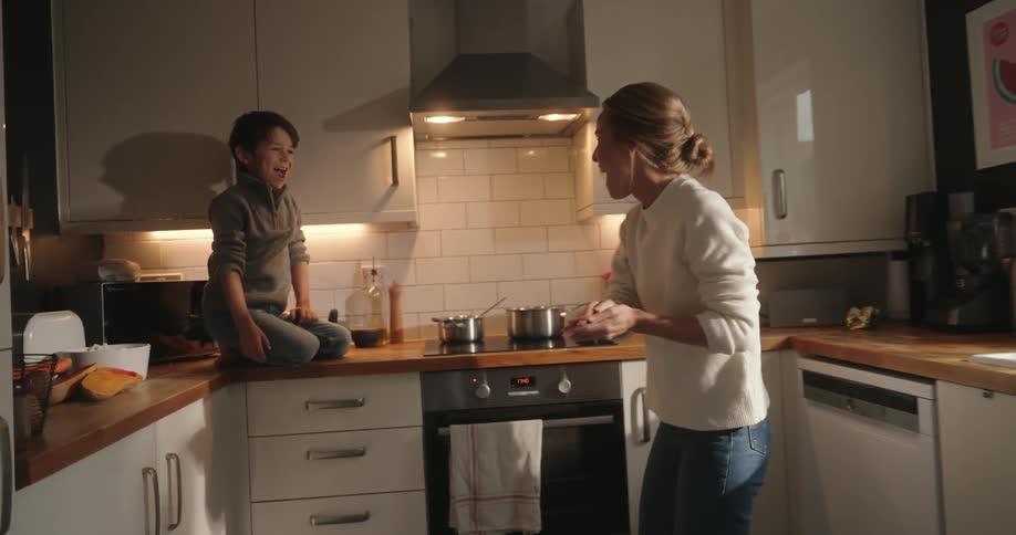 Mother playing with her little son who is on the kitchen counter throwing food