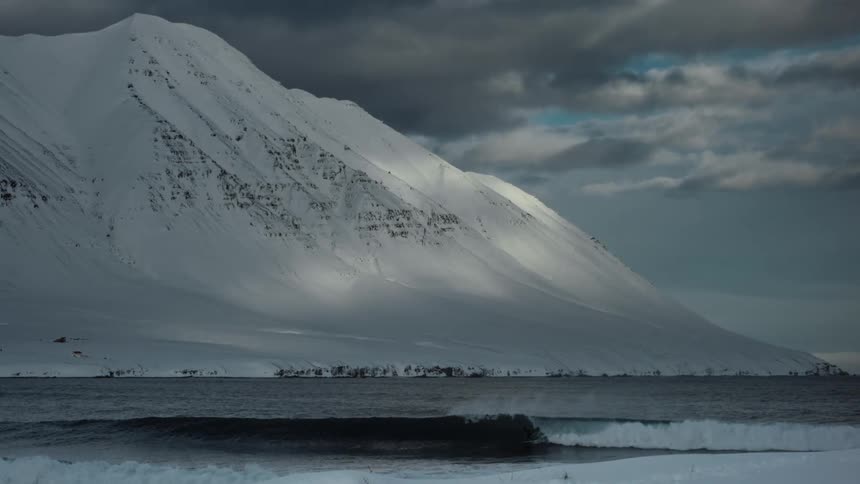 Surfer riding a wave on a beach at the edge of a snowcapped mountain on a foggy day
