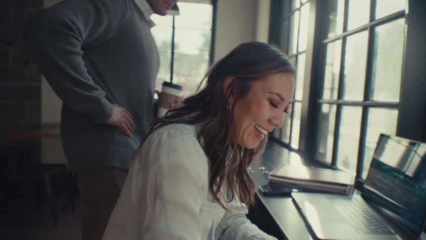 A woman in a white shirt is seated at a table working on a laptop and writing on a notebook