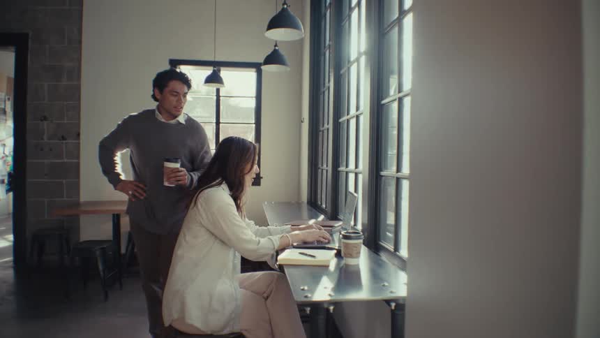 Man standing drinking coffee talking next to a woman who is sitting typing on a laptop