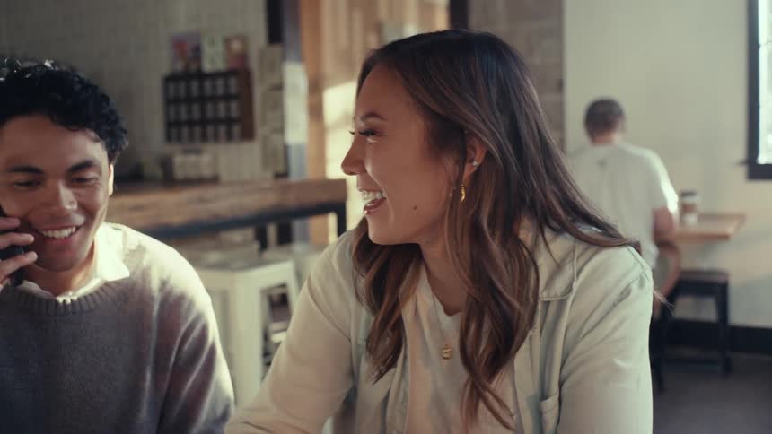 Two friends sitting at a table talking to one of them on a cell phone
