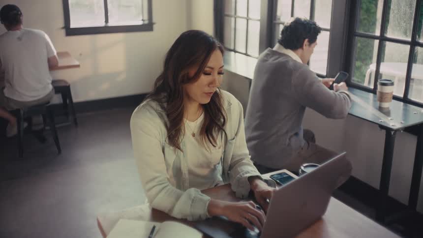 Three people in an open space office each with their cup of coffee working on smart phones or laptops with a woman drinking coffee then typing on her computer