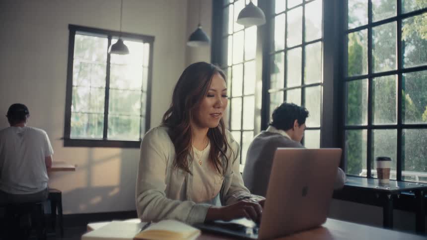 A woman sits at a table in a cafe typing on a laptop while two other people are in the background