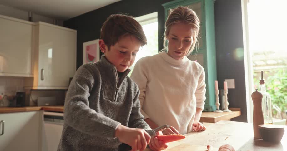 Mother watches son peeling carrots over the cutting board at home