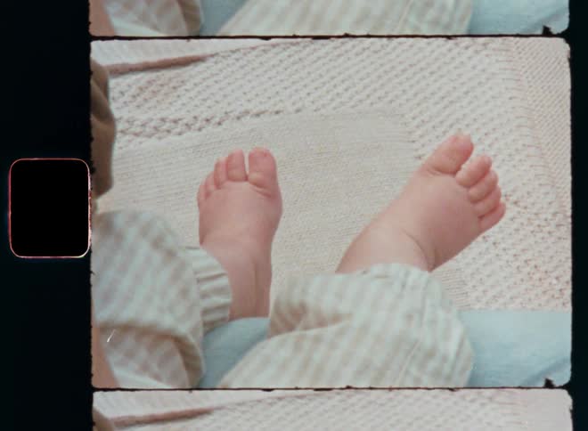 Super-8 close-up of the legs of an unrecognisable toddler or baby girl or boy relaxing in the arms of the caring mother while the child’s bare feet are dangling over a beige blanket outside on a sunny day in the garden