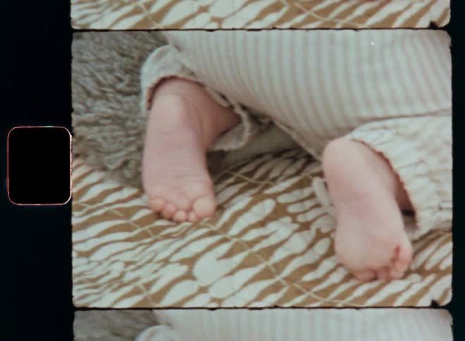 Hand-held Super 8mm close up of a crawling toddler’s bare feet wearing cute beige baby clothing while moving outside on a blanket in the garden on a sunny spring or summer day during early and happy childhood