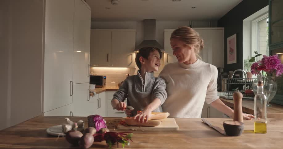 Mother in the kitchen giving a kiss to her son who is cooking a butternut squash