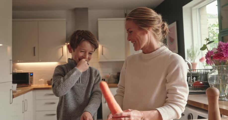 A blonde woman and a little boy are in a kitchen and are laughing happily while the woman holds a carrot and the little boy cleans his teeth using a toothpick