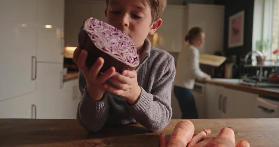 Boy holding a cabbage in the kitchen and his mother comes to his side