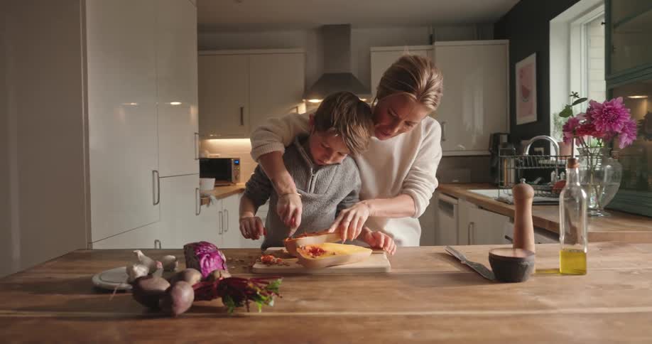 A mother helps her young son scrape seeds out of fruit on a cutting board in the kitchen