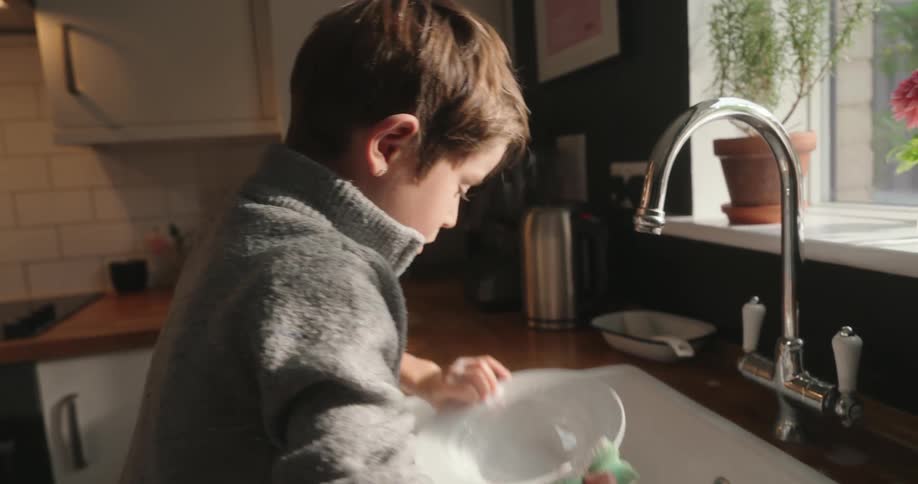 A young boy is washing a soup dish then tries to make a large soap bubble burst by blowing on it laughing when he fails
