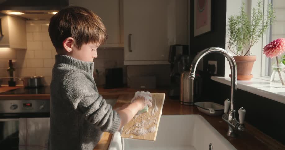 A young boy washes a small wooden cutting board with soap in a kitchen sink