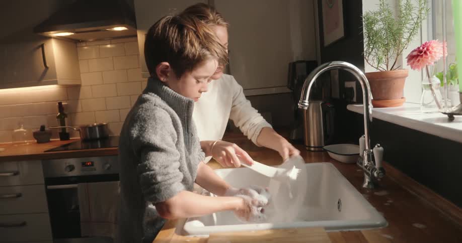 Mother and son stand at a kitchen sink one teaching the other how to wash dishes