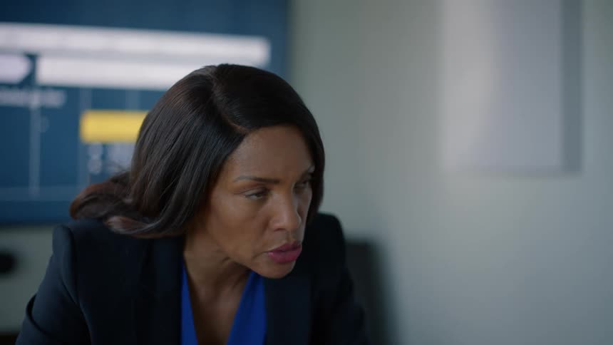 A woman wearing a black blazer and blue shirt is talking with someone standing in front of her