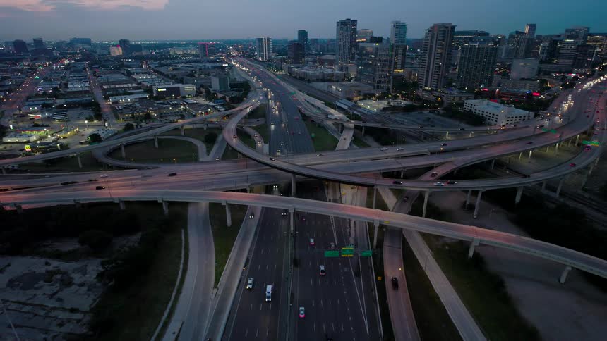 Aerial view of a multilevel highway interchange with numerous lanes and ramps during twilight with city buildings in the background