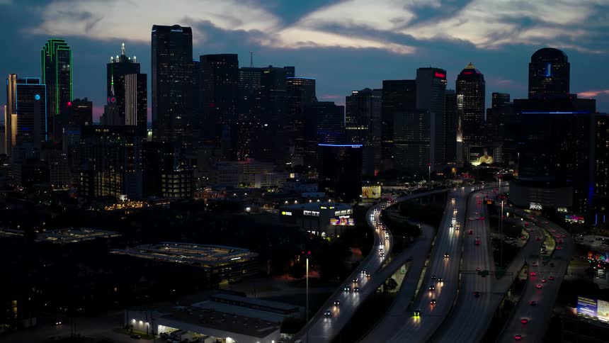 A sprawling cityscape at dusk illuminated by the headlights of cars on a freeway below