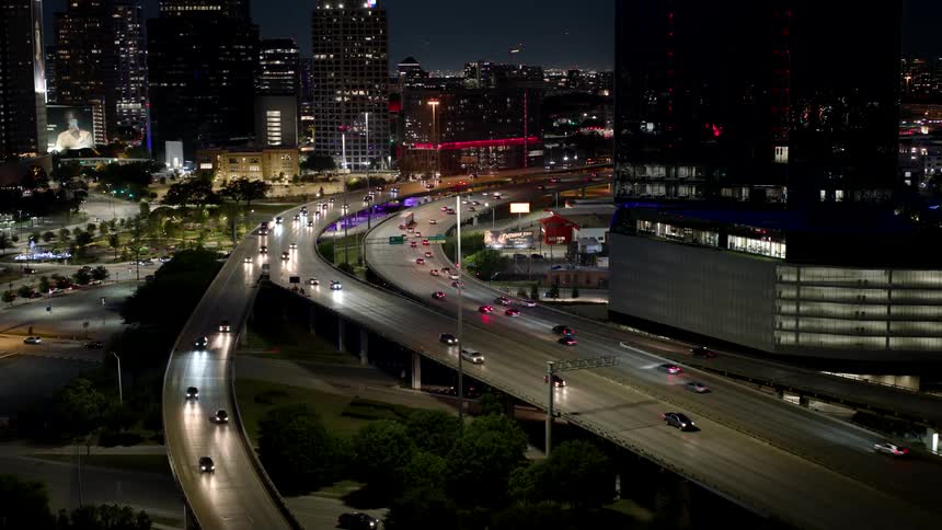 A busy highway curves through a city at night with cars traveling in both directions surrounded by illuminated buildings and the glow of streetlights