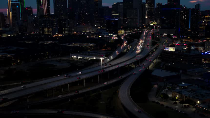 Nighttime aerial view of a multilevel road complex with vehicle lights creating trails along the curved roads surrounded by illuminated buildings