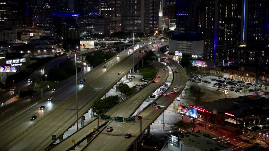 A big highway with several lanes and many cars passing by and there are several huge buildings around and it is night