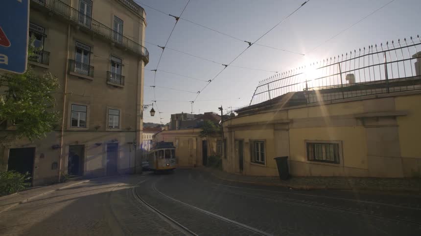 Yellow tram turns onto a curved street with buildings on either side and tram cables crossing overhead