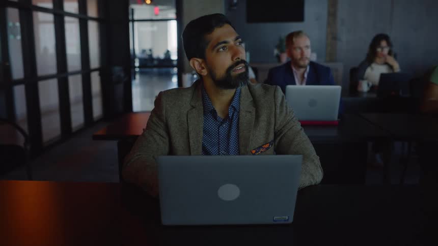 A dimly lit office space with a bearded man and several individuals working on laptops