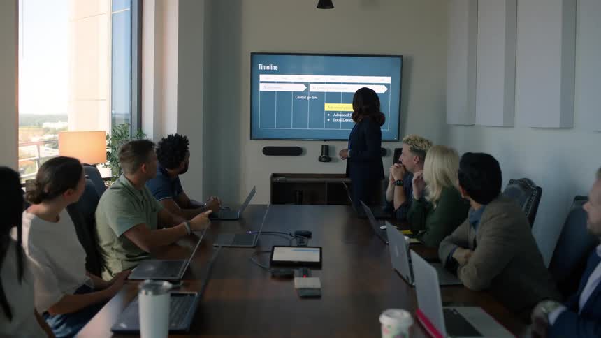 In a meeting room full of people sitting around a table with a laptop in front of each of them a woman standing presents a graph on a wall screen