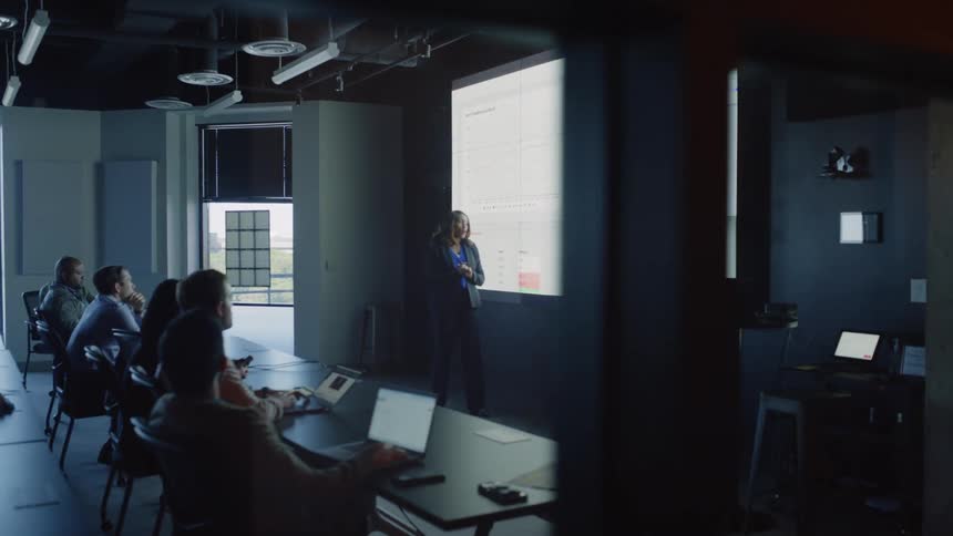 Person giving a presentation in a modern office meeting room with attendees seated and looking towards a large screen displaying a schedule or project timeline