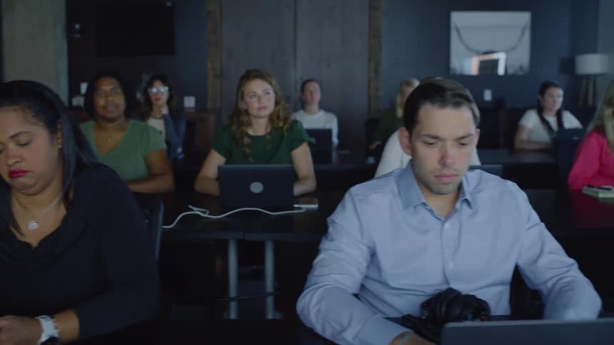 Several people sitting at black tables and are using laptops while looking straight ahead