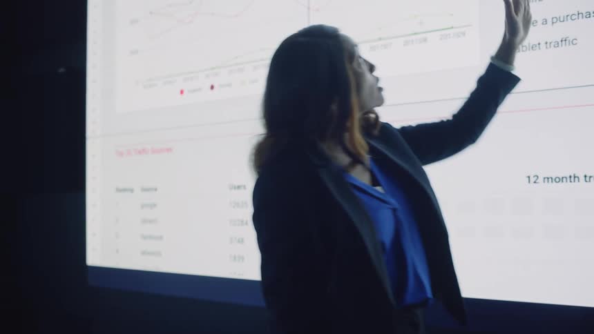 A woman is presenting in front of a projection of a chart to a room of people on laptops