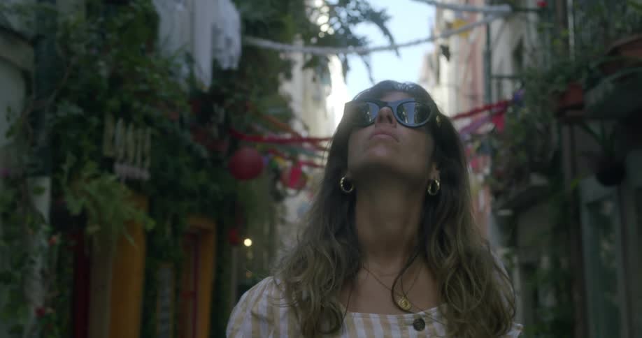 A woman is in an alley with plants on the facades of the houses on the sides and she is wearing sunglasses looking up