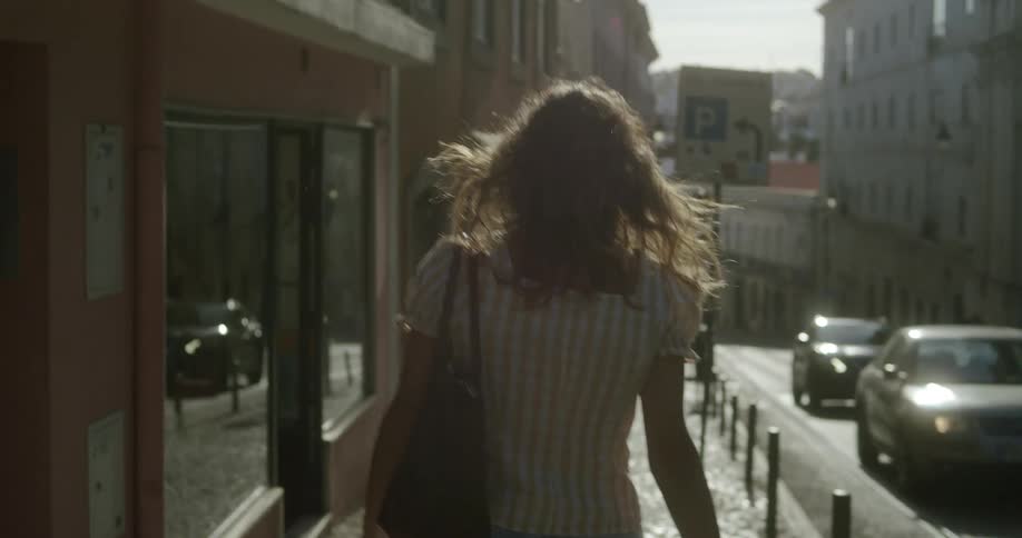 A woman is standing with her back turned holding a bag on her left shoulder and she is walking on a sidewalk and on the right side there are some cars passing on the street