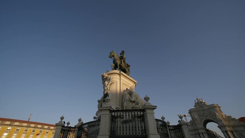 A statue of a man riding a horse on top of a white base surrounded by metal railings against a blue sky