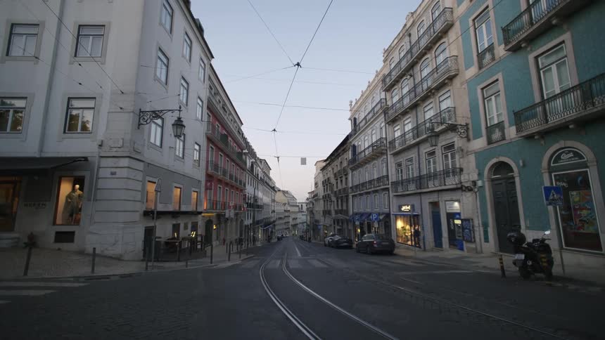 Street view with buildings on both sides tram lines above and vehicles parked along the curb